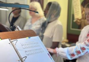 People singing at a choir with a music sheet in the foreground, featuring traditional hymns in a St. Mary Magdalene Church setting