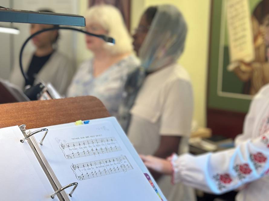 People singing at a choir with a music sheet in the foreground, featuring traditional hymns in a St. Mary Magdalene Church setting