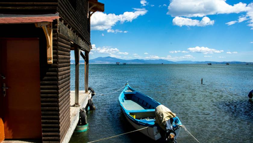 Blue boat tied to a wooden dock beside a rustic cabin overlooking a tranquil lake with a mountain backdrop