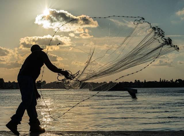 Silhouette of a fisherman casting a net at sunset over calm waters with a boat in the distance