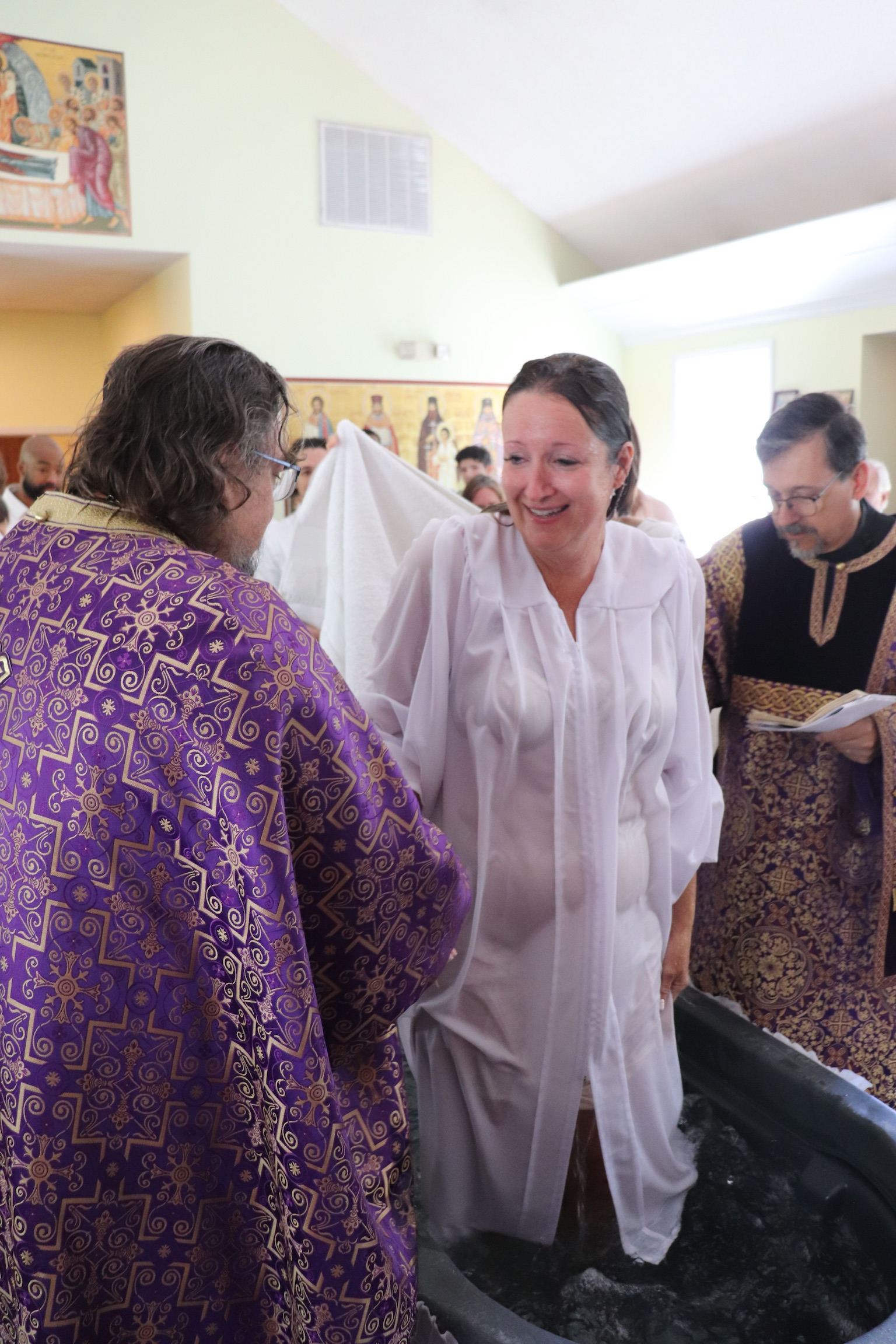 Woman in white robe getting baptized by priest in ornate purple robe in church setting