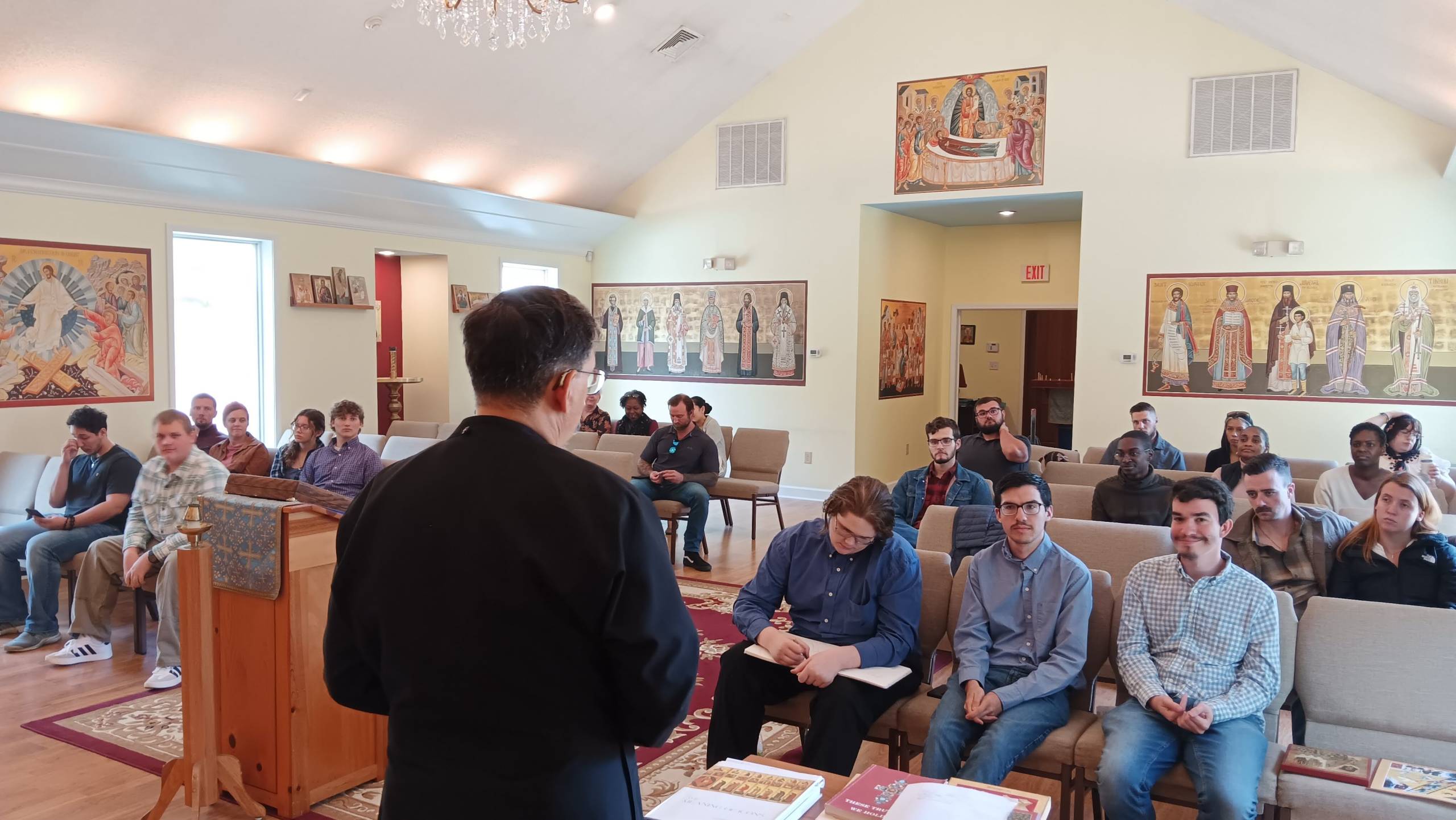 Man speaking to a seated group in a church room with religious art on the walls