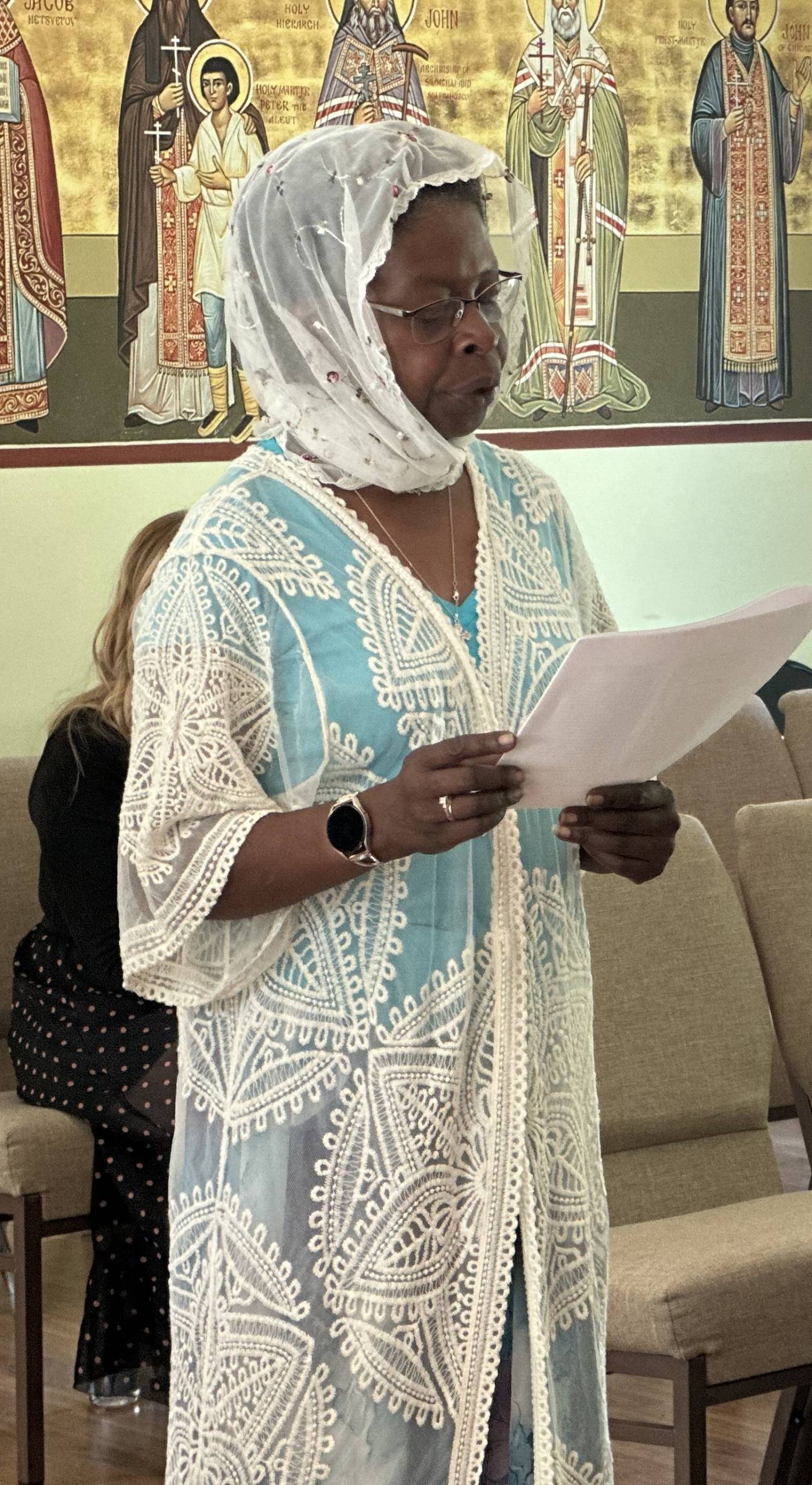 Woman in blue and white garment reading paper in front of religious artwork