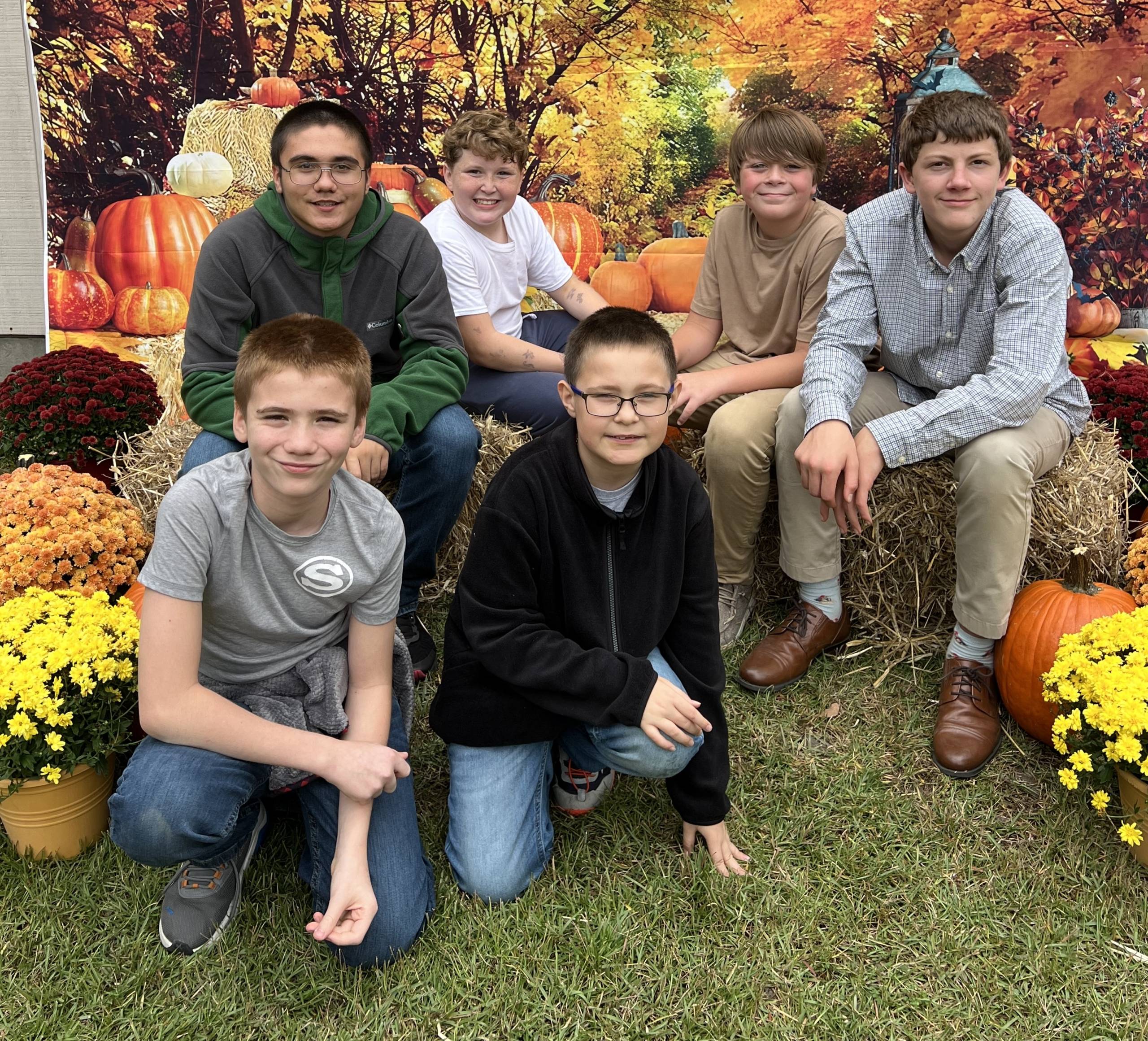 Group of kids sitting on hay bales surrounded by pumpkins and flowers in a fall-themed setting