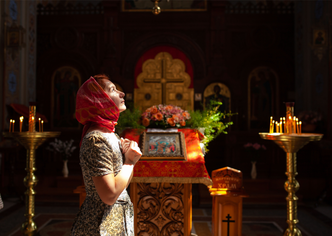 Woman in headscarf praying inside church near altar with lit candles and religious icons