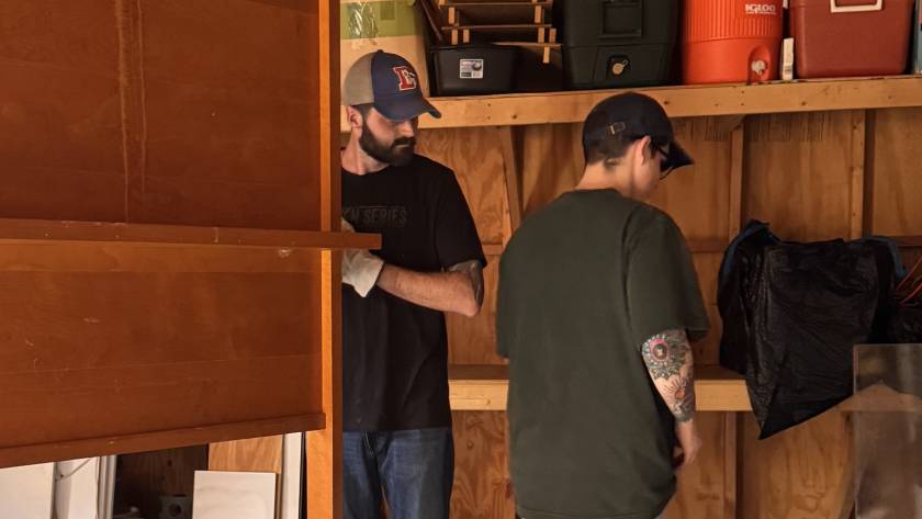Two men organizing tools in a wooden shed with shelves and storage containers