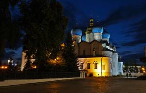 Illuminated cathedral with blue domes at night, surrounded by trees and a paved walkway