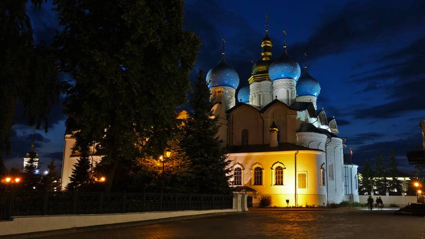 Illuminated cathedral with blue domes at night, surrounded by trees and a paved walkway