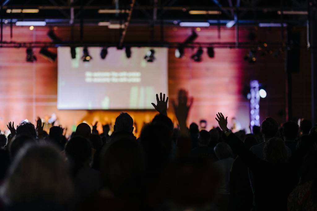 Audience raising hands at a conference with stage lights and presentation screen in background