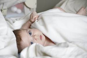 Baby lying under a white blanket, gazing with blue eyes on a cozy bed