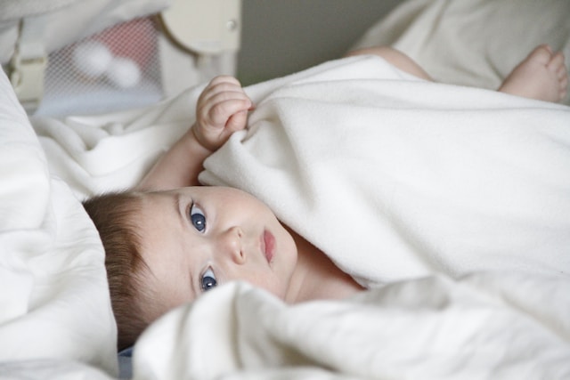 Baby lying under a white blanket, gazing with blue eyes on a cozy bed