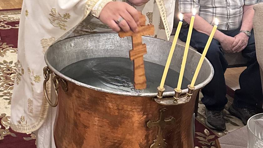 Priest immerses wooden cross in large baptismal basin during religious ceremony with lit candles nearby