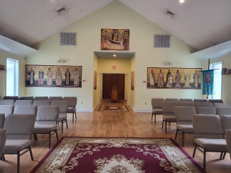 Church interior with rows of chairs, religious paintings on walls, and a red patterned carpet in the aisle