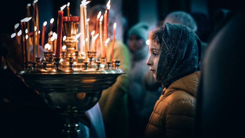 Child in a headscarf stands by lit candles during a solemn ceremony in dim lighting