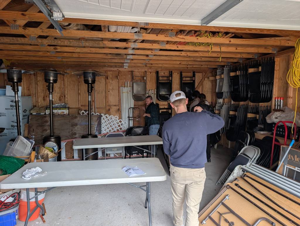 People organizing tables and chairs in a garage with wooden shelves and storage items visible