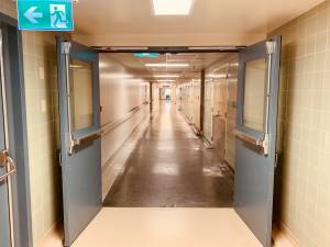 Open double doors leading to a long hospital corridor with emergency exit sign overhead