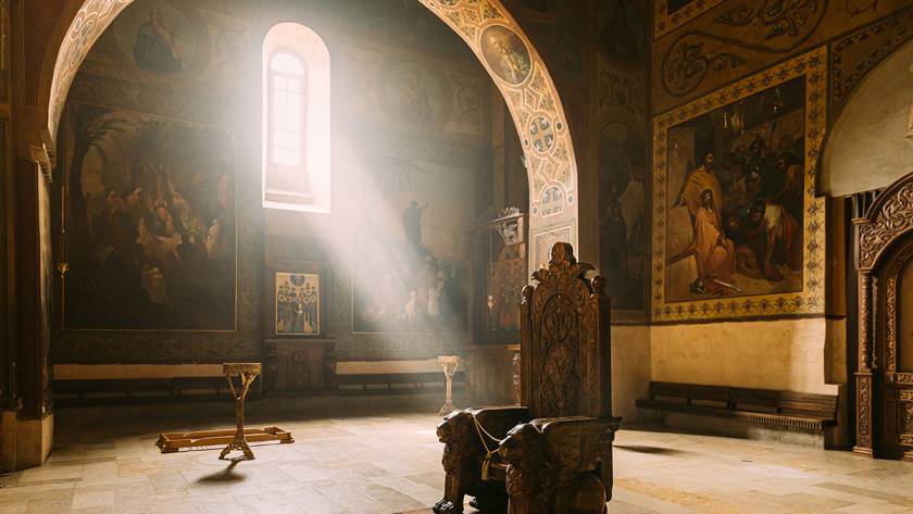 Ornate throne in a sunlit, historied church interior with decorative murals and arched window casting light rays.