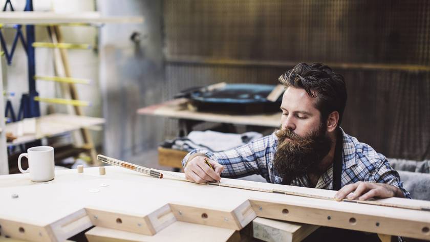 Bearded craftsman measuring wood in a workshop with a pencil and ruler, white mug on workbench
