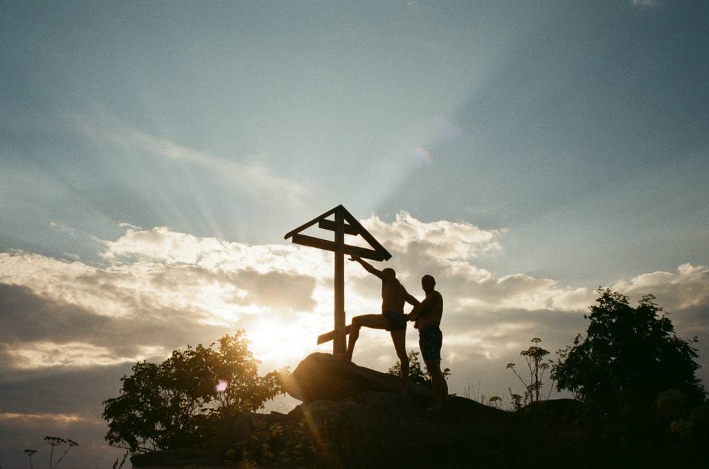 Silhouetted men by a wooden cross at sunset, standing on rocks, surrounded by trees and a dramatic sky.