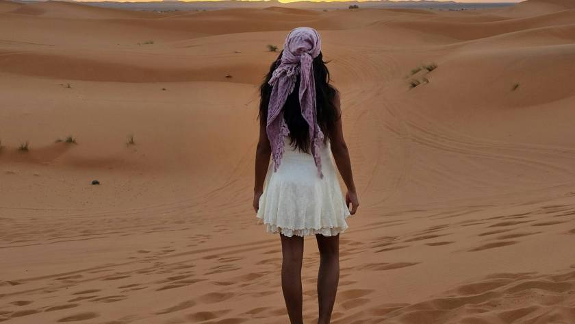 Woman in a white dress stands in sandy desert at sunset, facing the horizon, wearing a purple headscarf