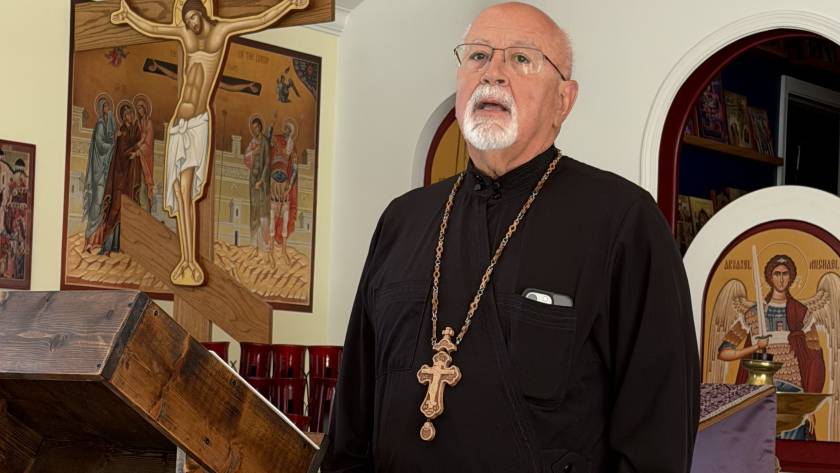 Elderly priest in black robe with cross necklace stands near religious icons in a church setting
