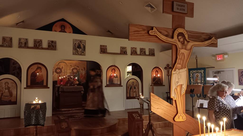 Orthodox church interior with icons, large crucifix, and candles during a service with people and priest in attendance
