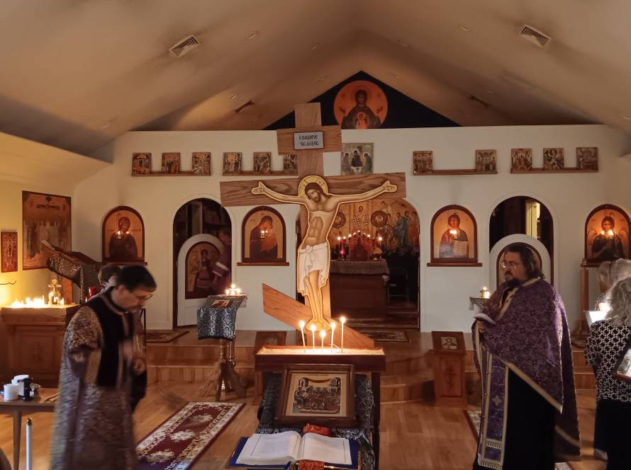 Orthodox church interior with icons, cross, and candles during a service with clergy and congregation members.