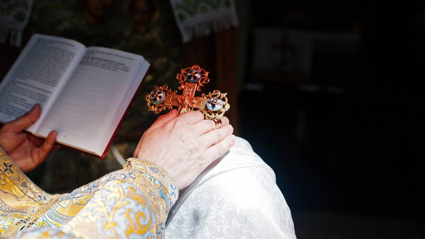 Ornate cross held by a priest in embroidered vestments, with an open book in dimly lit church setting