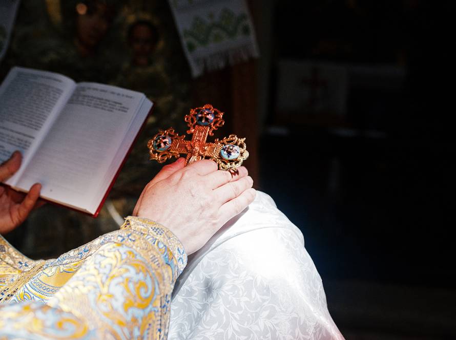 Ornate cross held by a priest in embroidered vestments, with an open book in dimly lit church setting