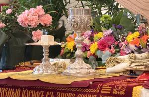 Ornate chalice and plate with bread on floral-decorated altar setting, vibrant flowers in the background