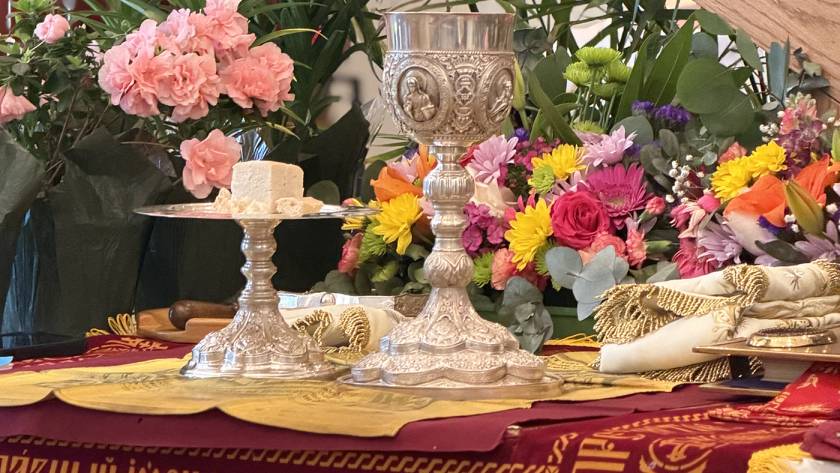Ornate chalice and plate with bread on floral-decorated altar setting, vibrant flowers in the background