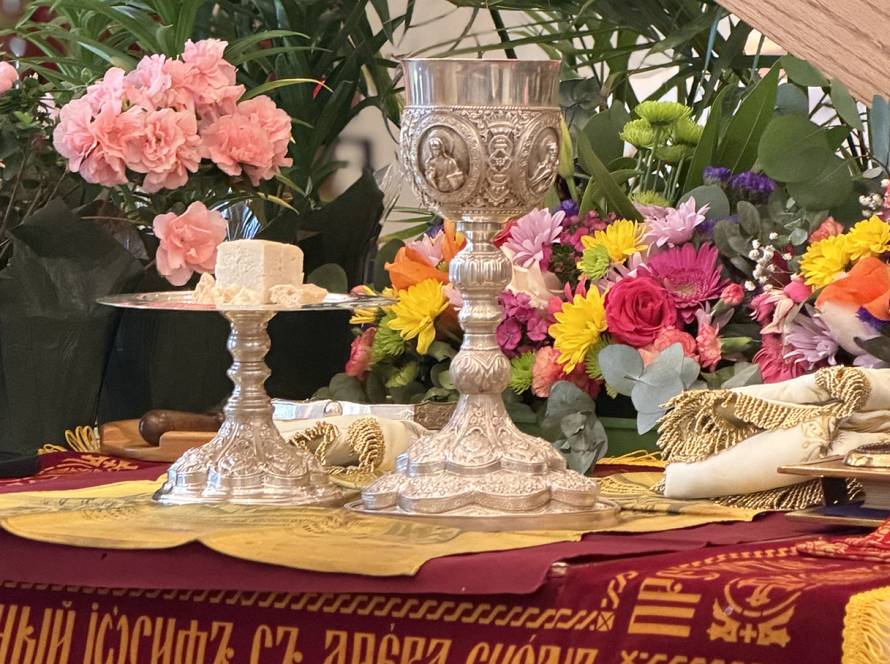 Ornate chalice and plate with bread on floral-decorated altar setting, vibrant flowers in the background