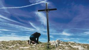 Person kneeling by a cross on a hilltop with a vibrant blue sky and clouds in the background