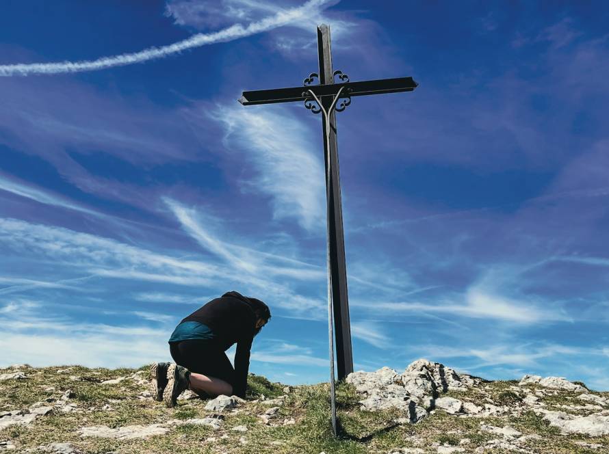 Person kneeling by a cross on a hilltop with a vibrant blue sky and clouds in the background