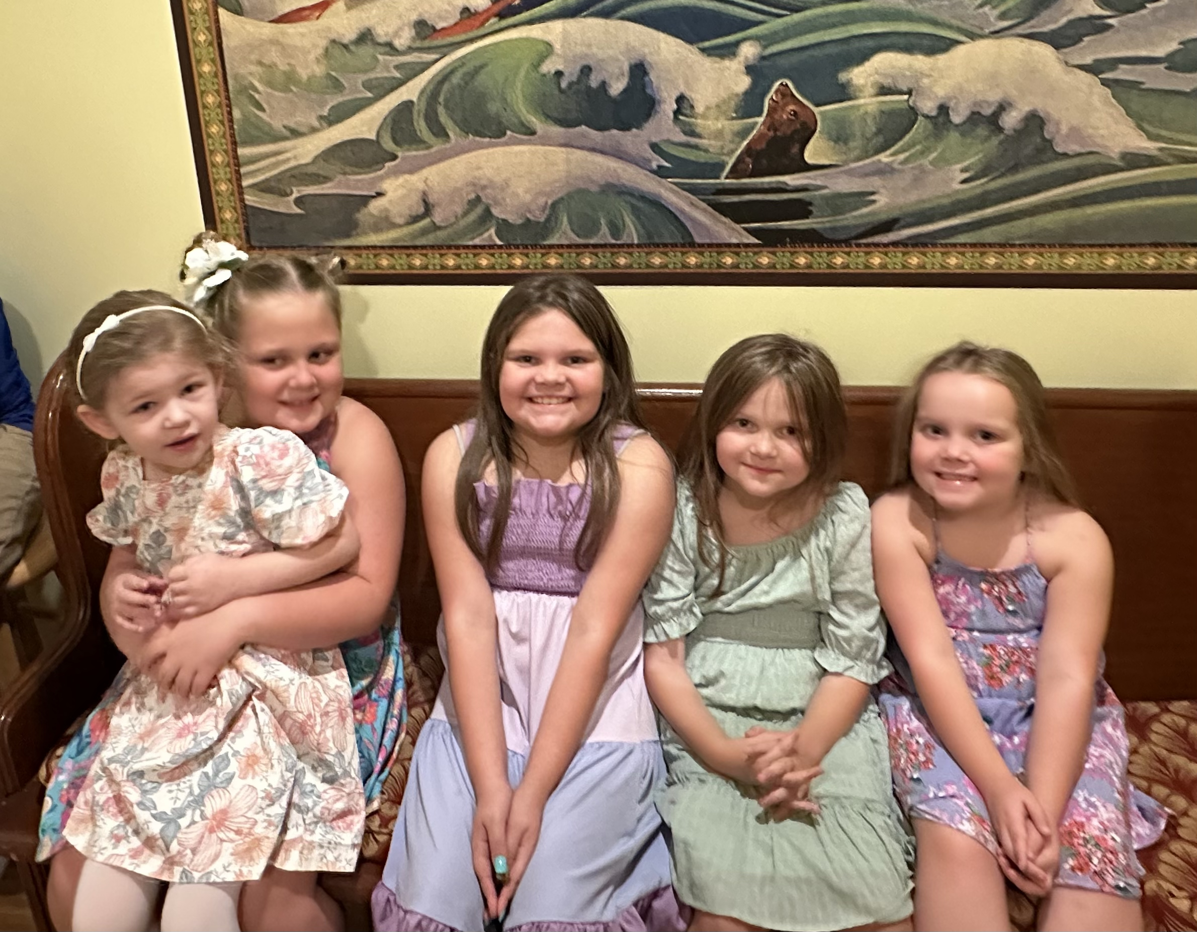 Five smiling children sitting on a bench in colorful dresses with a vibrant ocean-themed painting in the background