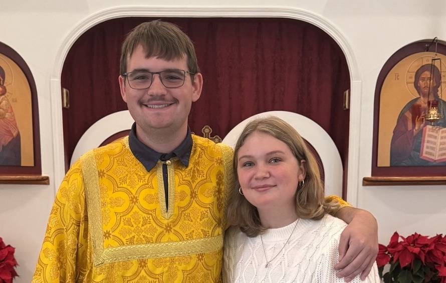 Two smiling people in a church setting with religious icons and decorative red poinsettias
