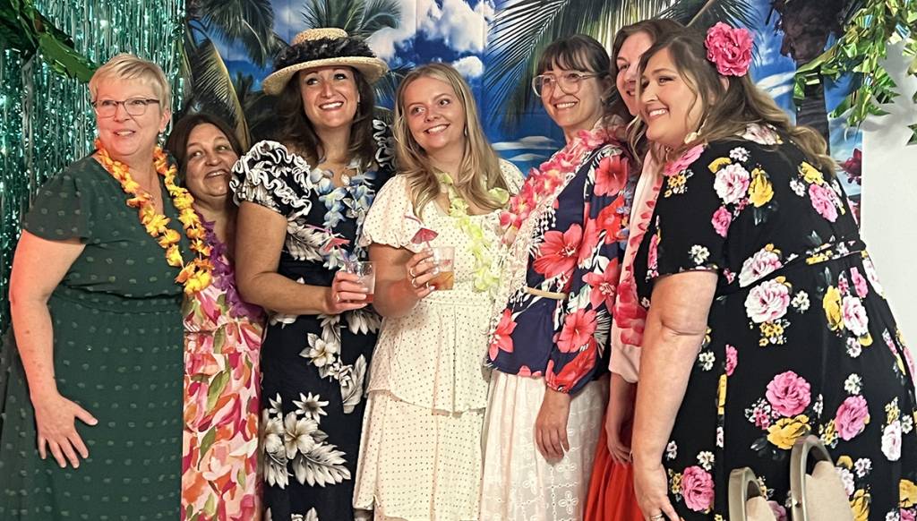 Group of women in Hawaiian-themed outfits smiling at a tropical party setup with floral decorations and leis