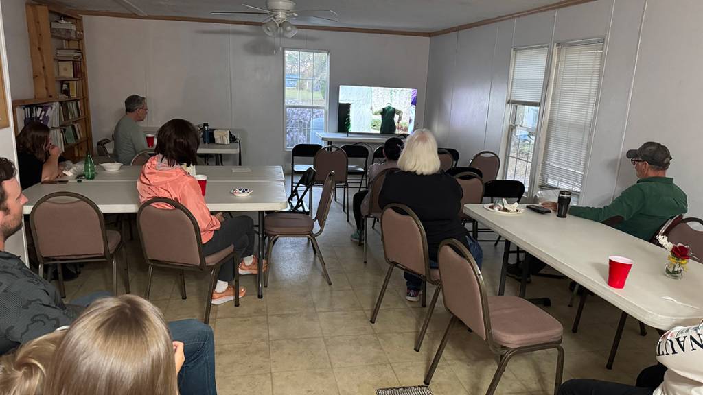 People watching TV in a community room with chairs and tables, snacks on tables, and a small flat screen TV in the background