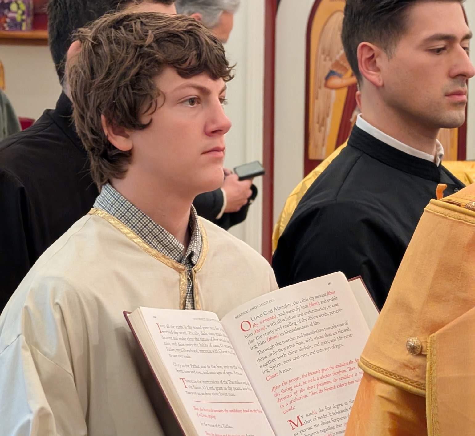 Young man in religious attire holding an open book during a church service, standing attentively among others