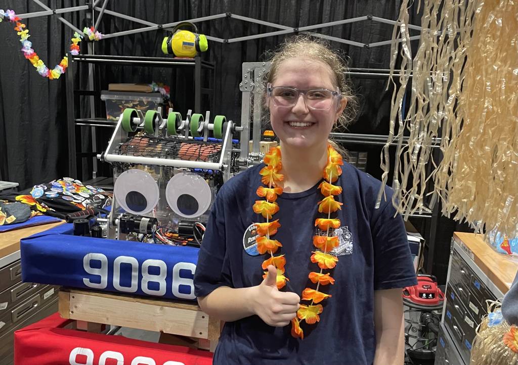 Smiling participant at a robotics competition with a robot and colorful accessories in the background