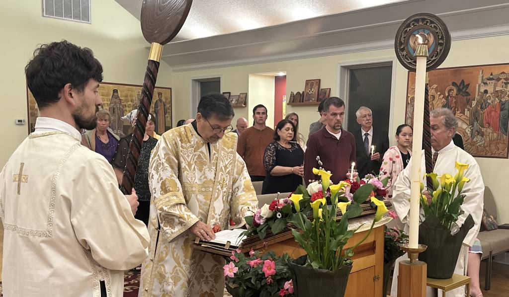 Orthodox Easter service with priest reading, surrounded by people holding candles and floral decorations