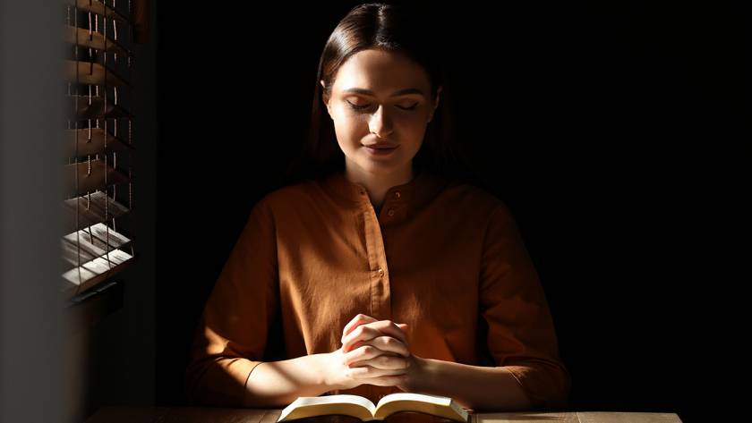 Woman in brown shirt sitting at wooden table, hands clasped in prayer, beside an open book, in dimly lit room