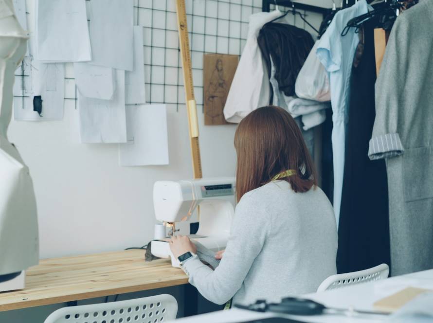 Seamstress working on a sewing machine in a design studio with clothing patterns and garments hanging nearby