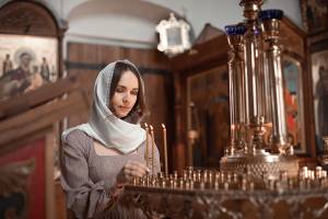 Woman lighting candles in a church, wearing a white headscarf, surrounded by religious icons and ornate decorations