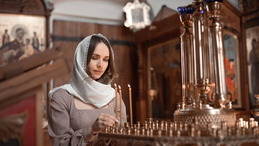 Woman lighting candles in a church, wearing a white headscarf, surrounded by religious icons and ornate decorations