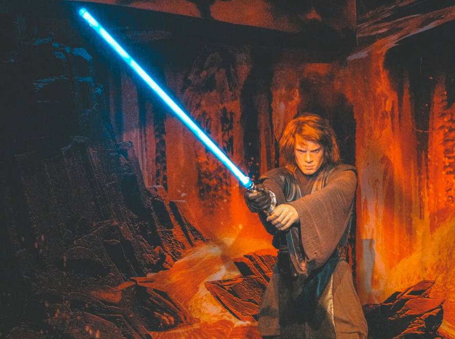 Person wielding blue lightsaber in dramatic, fiery setting with rocky backdrop and intense expression