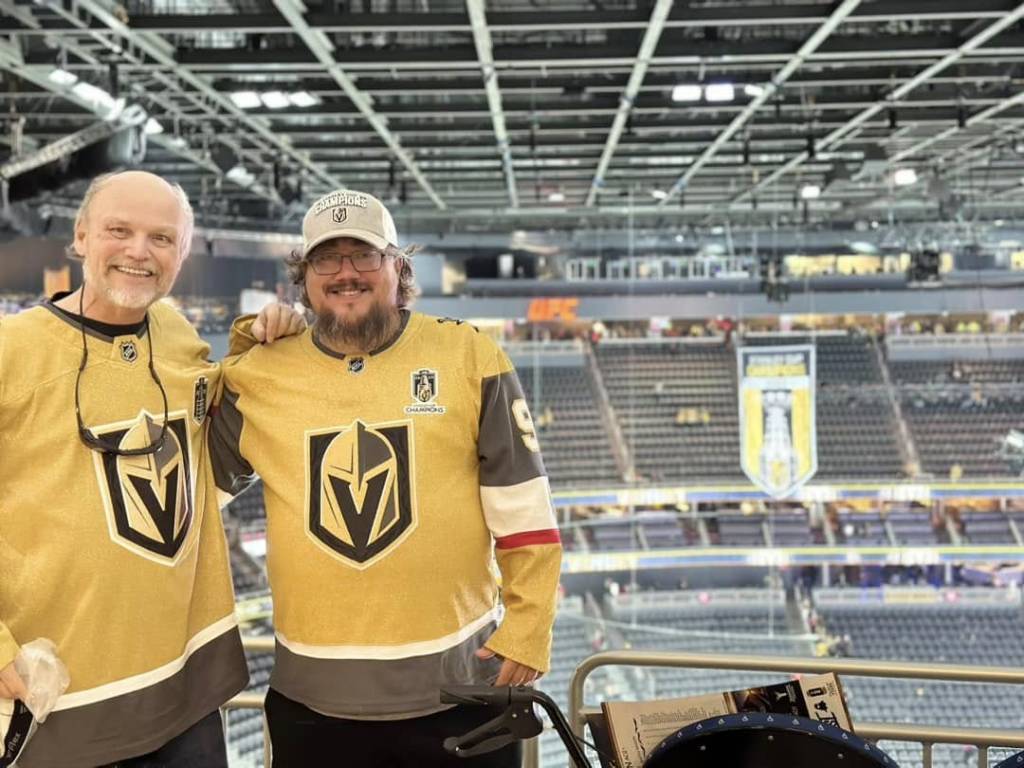 Two hockey fans in Vegas Golden Knights jerseys at an arena with empty seats in the background.
