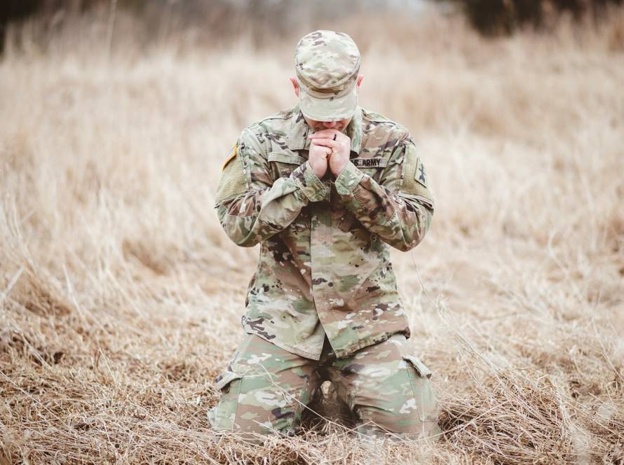Soldier kneeling in a field, wearing camouflage uniform, with hands clasped in thought or prayer