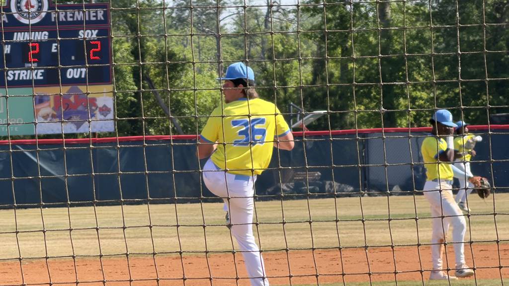 Baseball pitcher in yellow jersey preparing to throw, scoreboard showing 2-2 score in outdoor game setting