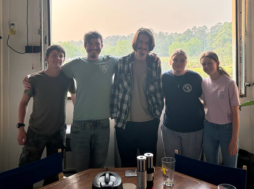Group of five young people smiling indoors by a large window with trees in the background
